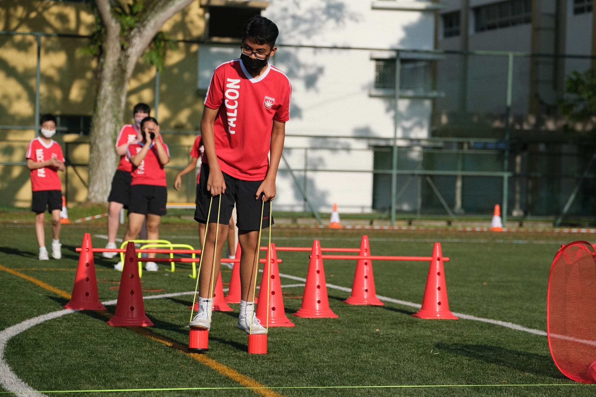 A masked boy in a red Falcon jersey navigates an obstacle course with red cones using elastic bands on a sports field.